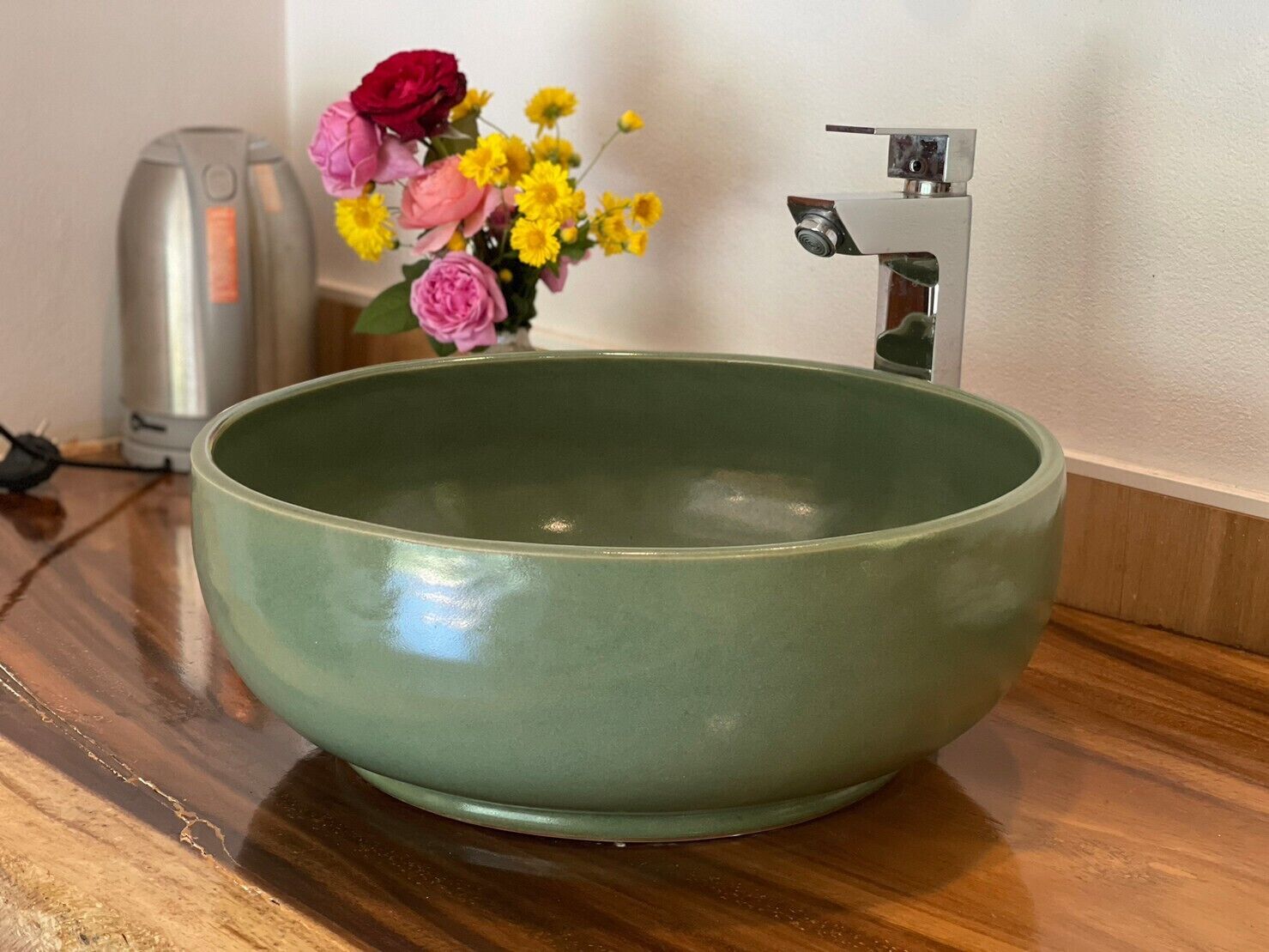 A green ceramic sink with a modern faucet, a kettle, and a vase of flowers on a wooden countertop.