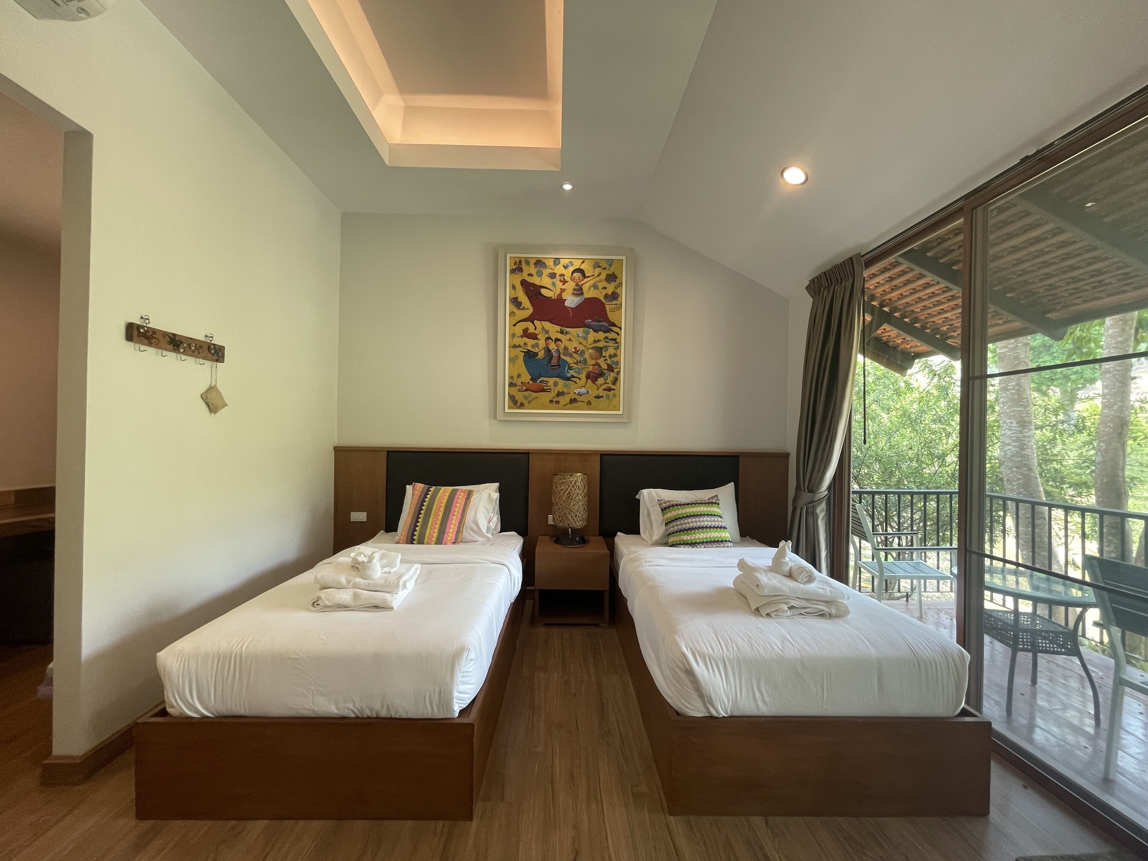 Une chambre d’hôtel avec deux lits, une grande fenêtre donnant sur des arbres, un luminaire au plafond et un tableau accroché au mur.