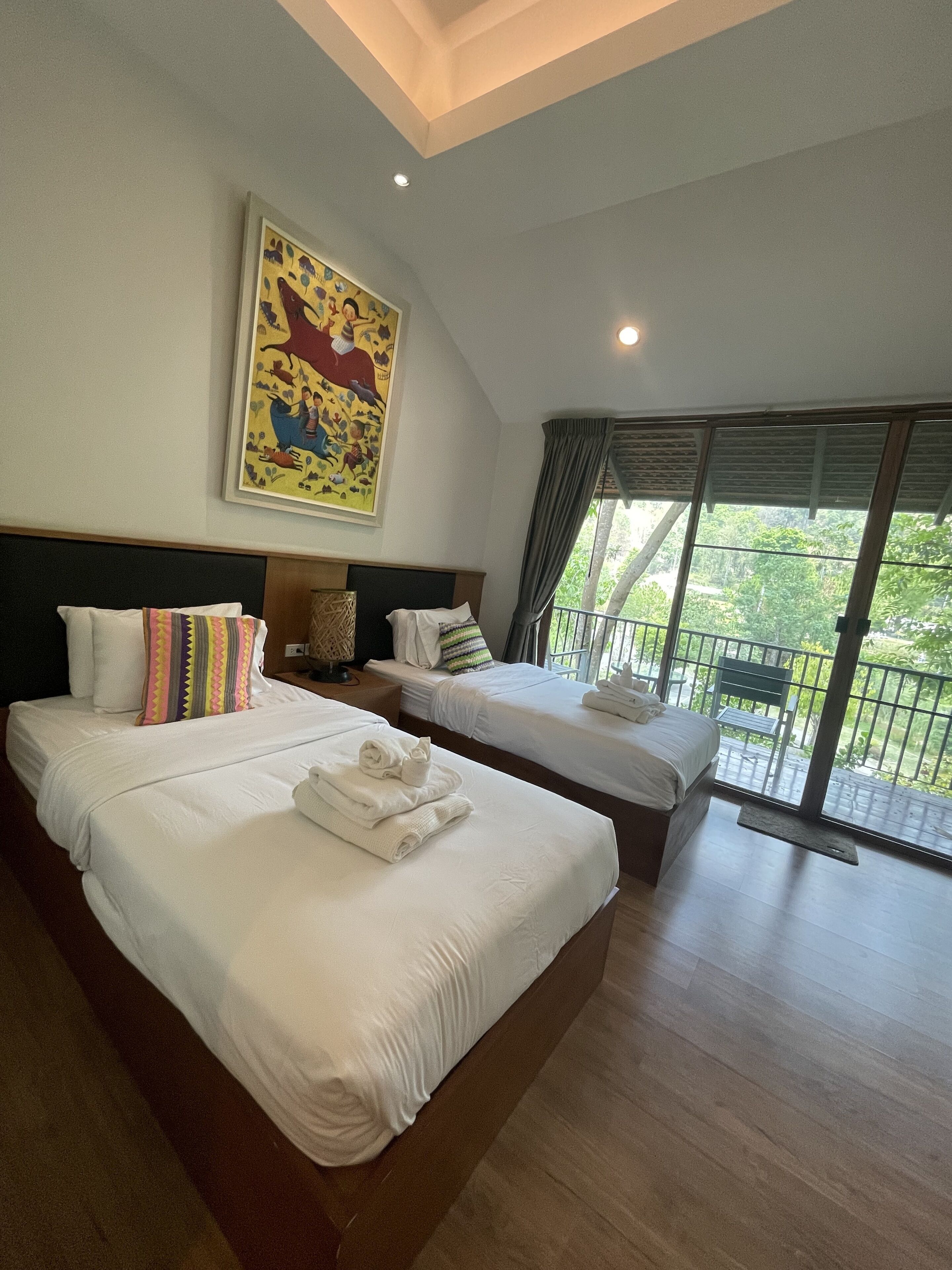 A hotel room with two beds, a large painting, and a balcony with a view of greenery.