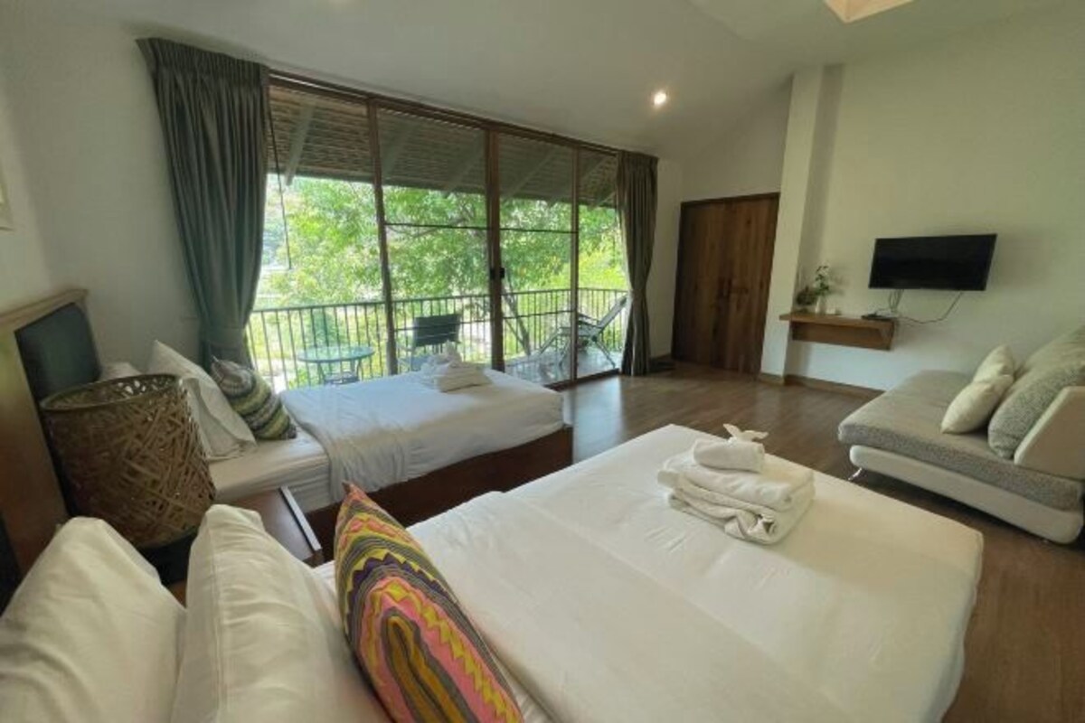 A hotel room with a large bed, a sofa, a TV, and a balcony with a view of trees.