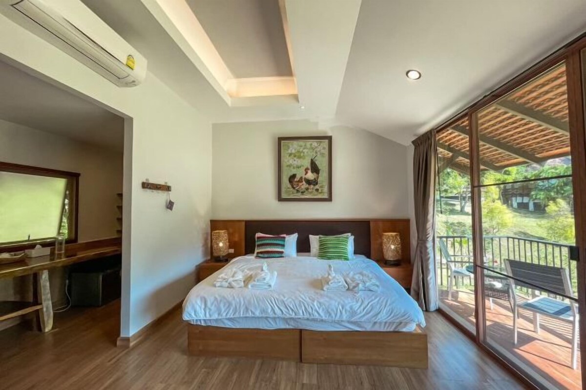 A bedroom with a large bed, a TV, and a balcony with a view of greenery.
