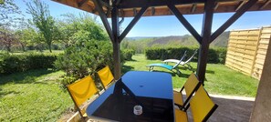 A covered patio with a table and chairs, overlooking a grassy area with trees and a wooden fence.