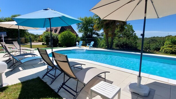 En swimmingpool med solstolar och parasoller, omgiven av grönska och ett hus i bakgrunden.