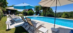 Une piscine entourée de chaises longues et de parasols, avec de la verdure tout autour et une maison en arrière-plan.
