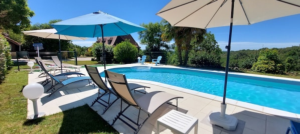 A swimming pool with lounge chairs and umbrellas, surrounded by greenery and a house in the background.