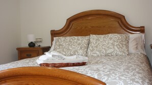 Un lit bien fait, avec une tête de lit en bois, une table de chevet sur laquelle se trouve une lampe, et un panier rempli de serviettes pliées.