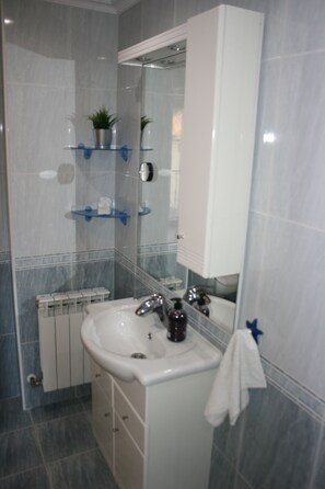 Ein Badezimmer mit einem weißen Waschbecken, einem Spiegel darüber und einem Schrank darunter. Über dem Waschbecken befinden sich zwei Ablagen, eine mit einer Pflanze und die andere mit einer Flasche.