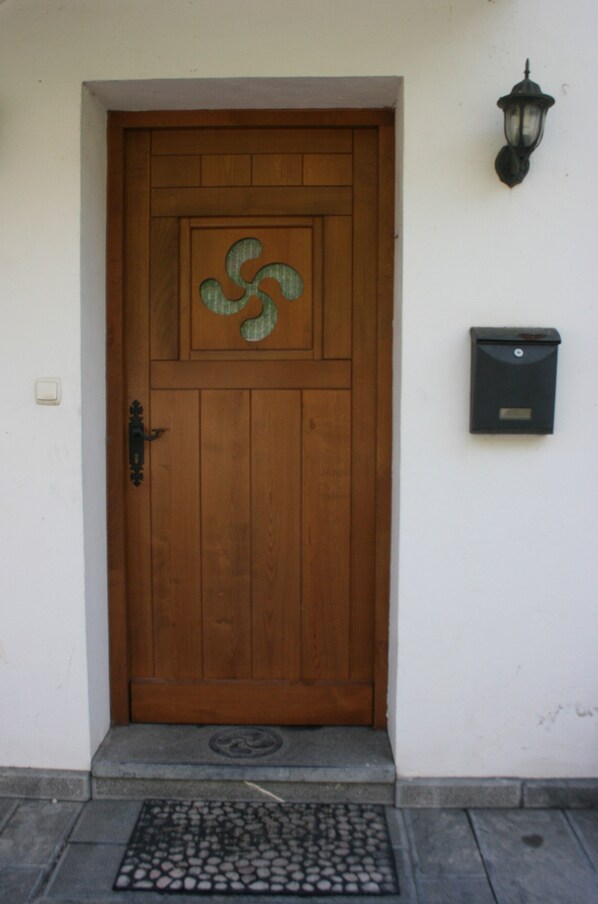 Une porte en bois ornée d’un motif métallique décoratif, une boîte aux lettres et un luminaire fixé au mur.