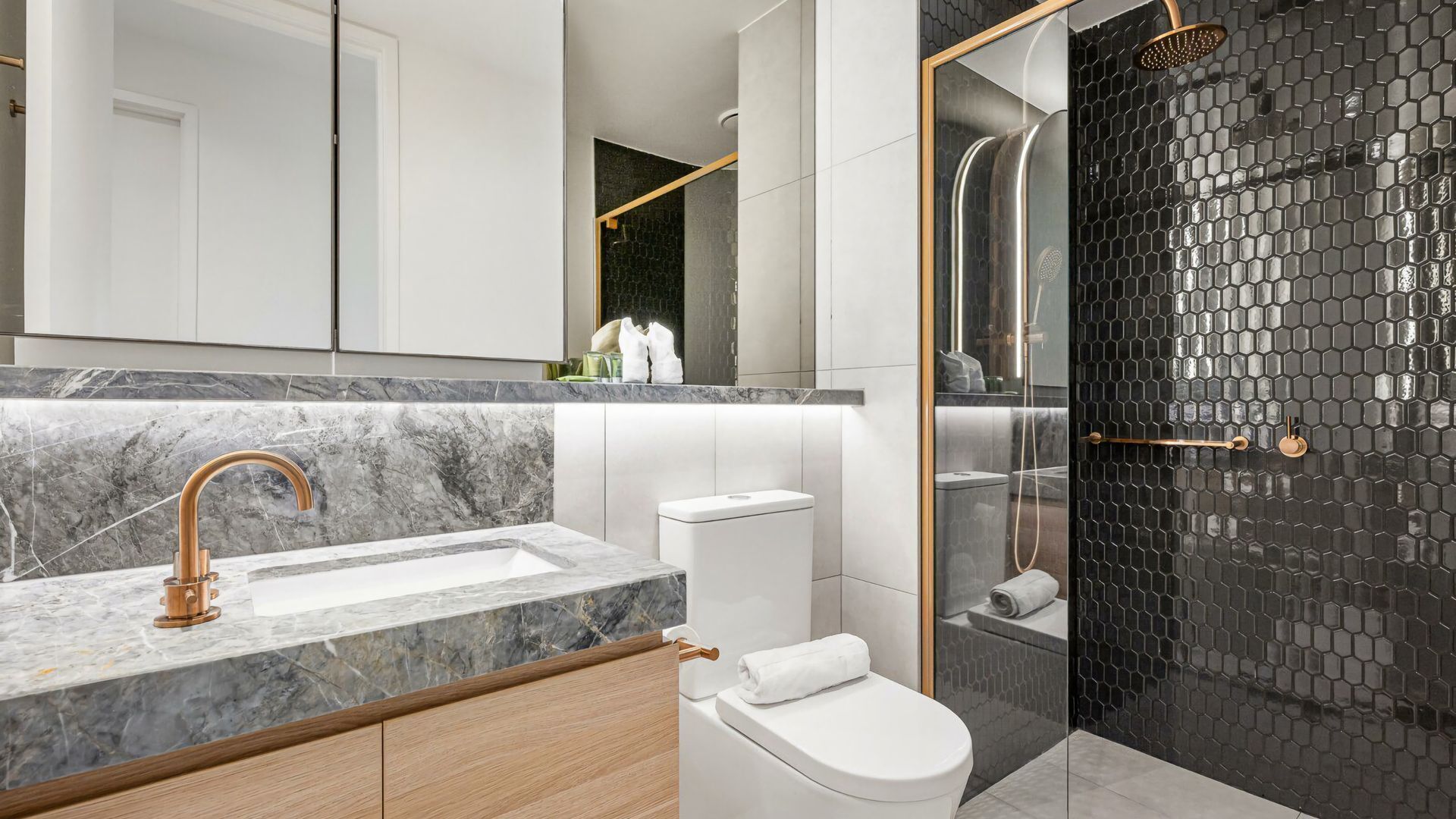 A modern bathroom with a marble sink, a toilet, and a shower with a black hexagonal tile wall.