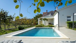 A rectangular swimming pool with a white border, surrounded by a paved patio. There are two lounge chairs with grey cushions.