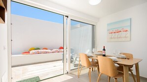 Un'area pranzo con un tavolo apparecchiato per due, una porta scorrevole in vetro che dà su un balcone con cuscini colorati e un quadro di fenicotteri sulla parete.