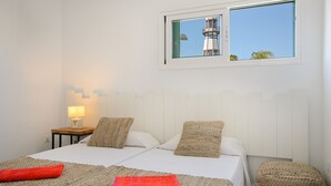 Una cama doble con dos almohadas, una mesita de noche con lámpara, una ventana con vista a un faro y un reposapiés tejido.