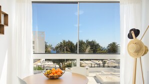 Un comedor con mesa y sillas, un bol de fruta, y vistas a un edificio y palmeras a través de amplios ventanales.
