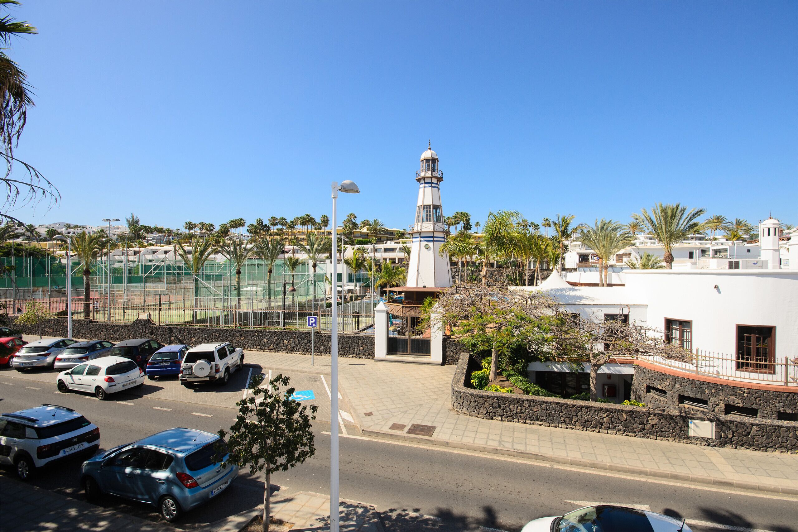 A coastal road with parked cars, a lighthouse, and buildings with balconies.