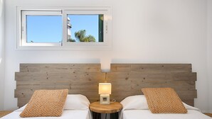 A double bed with a wooden headboard, two pillows, a bedside table with a lamp, and a window with a view of buildings and trees. - Blue Volcano • Pool and Gym • Beach 1 min • LVH (Puerto del Carmen)