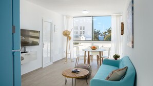 A modern living room with a blue sofa, a round coffee table, and a TV. There's a balcony with a view of palm trees and buildings. - Blue Volcano • Pool and Gym • Beach 1 min • LVH (Puerto del Carmen)