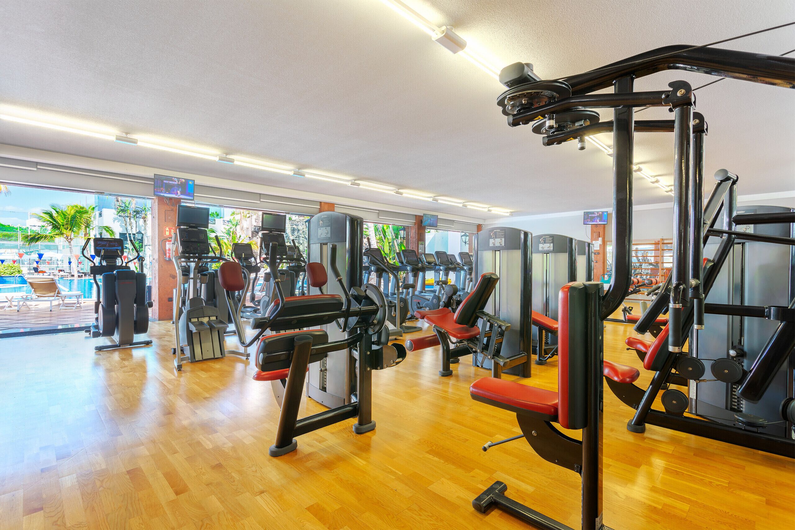 Een sportschool met diverse fitnessapparaten, een houten vloer en grote ramen met uitzicht op palmbomen en een zwembad.