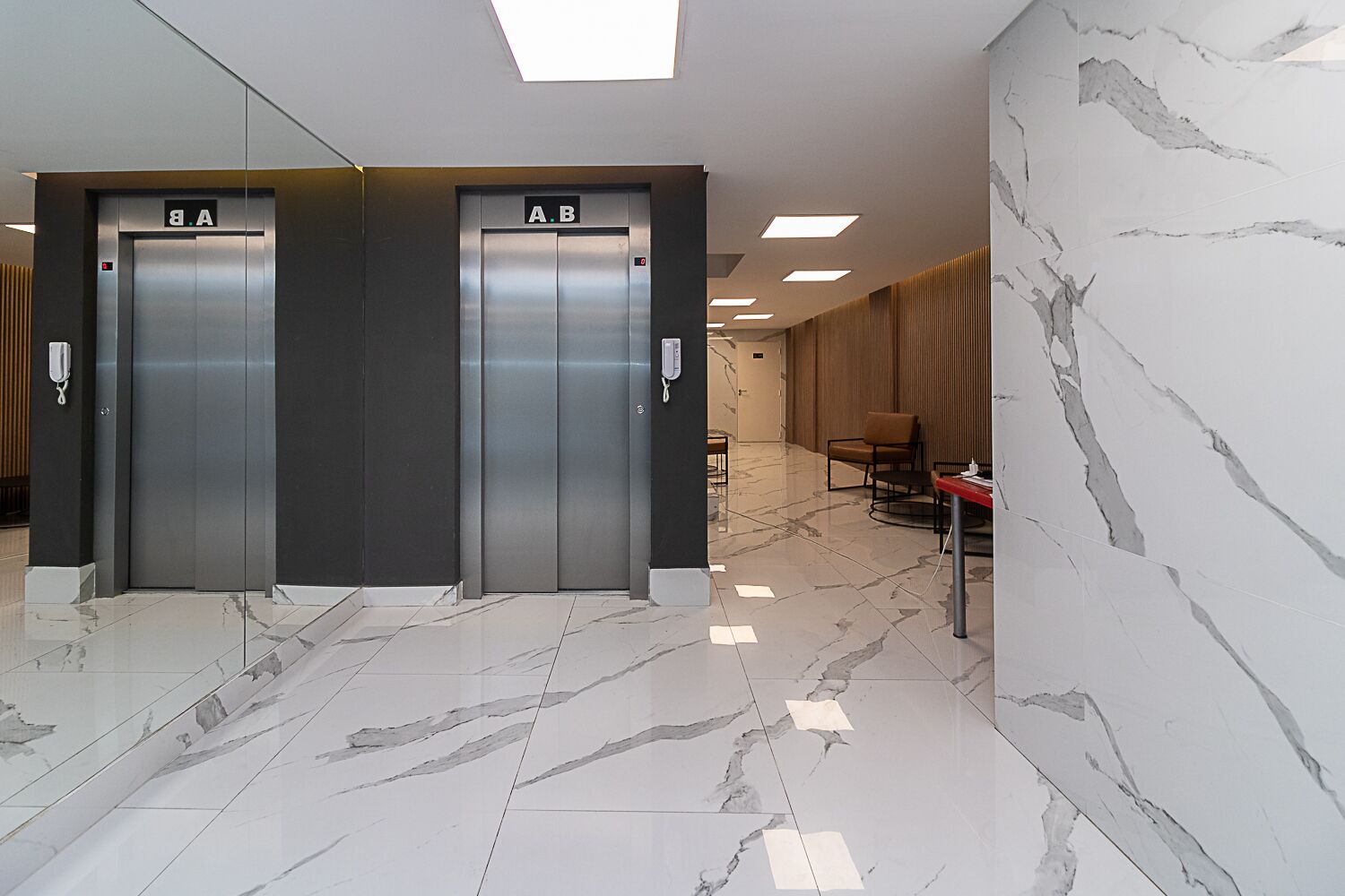Two elevator doors labeled A and B, a marble floor, and a wooden wall with a red table and chairs.