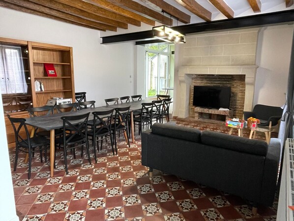A living room with a fireplace, a sofa, a dining table with chairs, and a bookshelf. - Spacious Family Cottage with Garden, Terrace, and Wifi near Le Mans and Tours (Chahaignes)