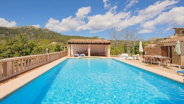 A clear blue swimming pool with a wooden deck, a covered seating area, and a mountainous backdrop.