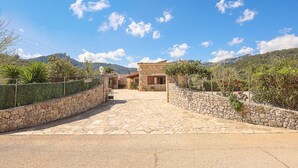 A stone pathway leading to a rustic house with a stone wall on one side and a fence on the other, surrounded by greenery and hills.