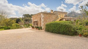 A stone house with a gravel driveway, surrounded by greenery and a mountain in the background.