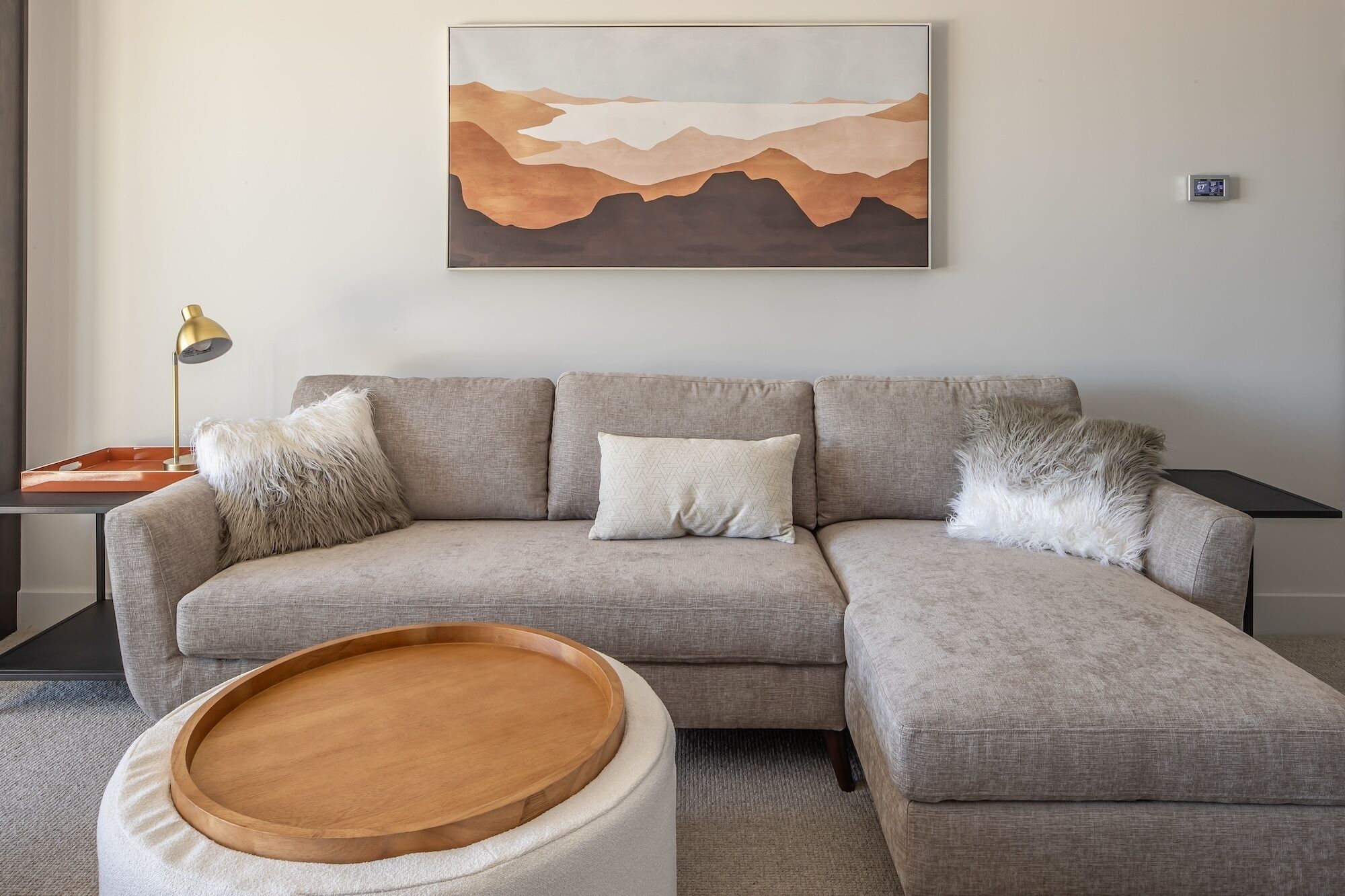 Ein modernes Wohnzimmer mit einem grauen Ecksofa, einem runden Holztisch und einem flauschigen weißen Pouf. Ein Gemälde von Bergen hängt an der Wand.