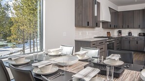 A modern kitchen with dark wood cabinets, a large window with a view of trees and mountains, and a dining area with a glass table set for four.