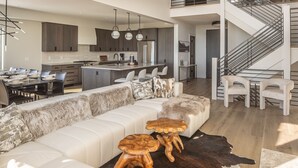 A modern open-plan living area with a sofa, two wooden stools, a dining table, and a kitchen in the background.