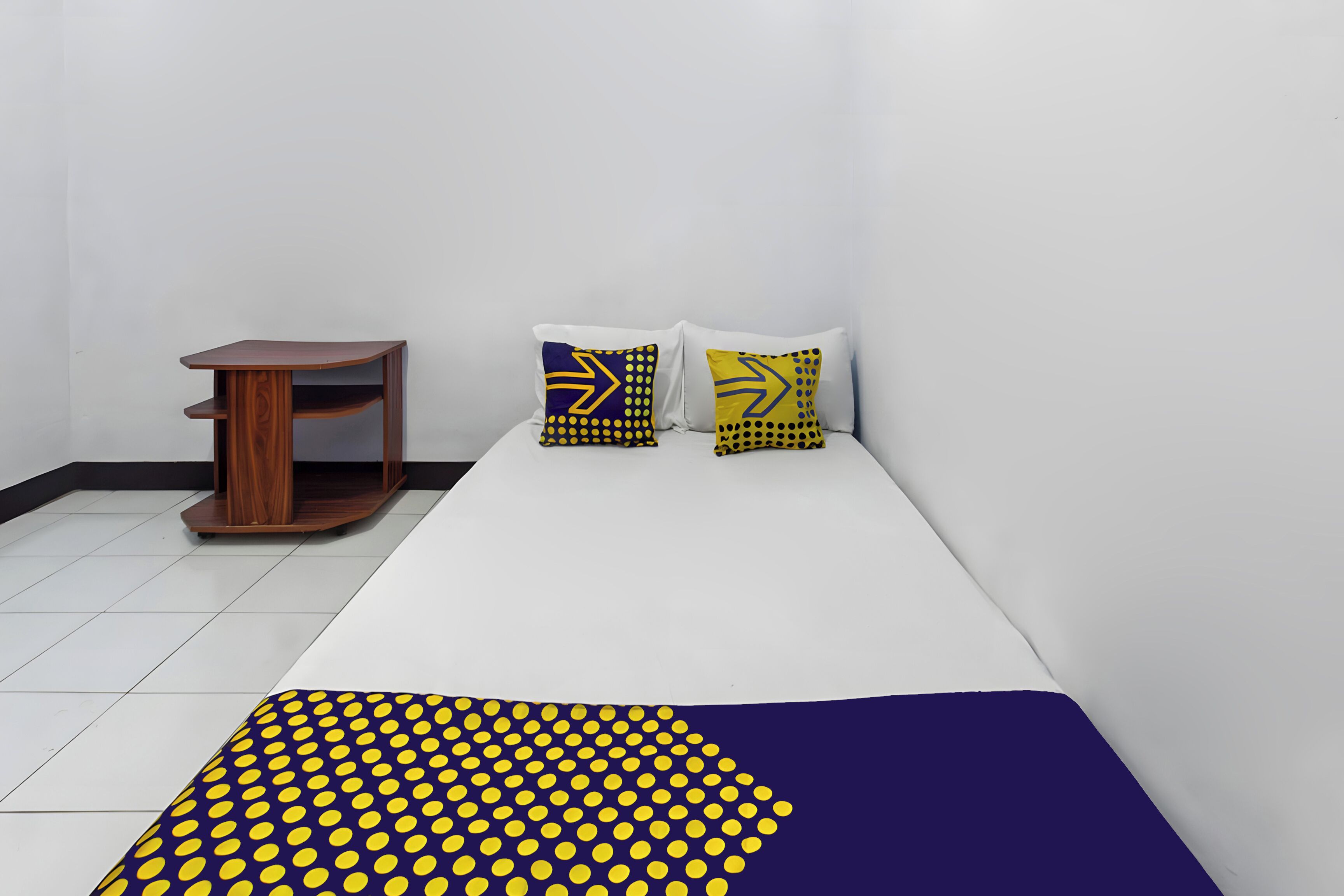 A room with a bed, a small wooden side table, and two pillows with a yellow and blue pattern.