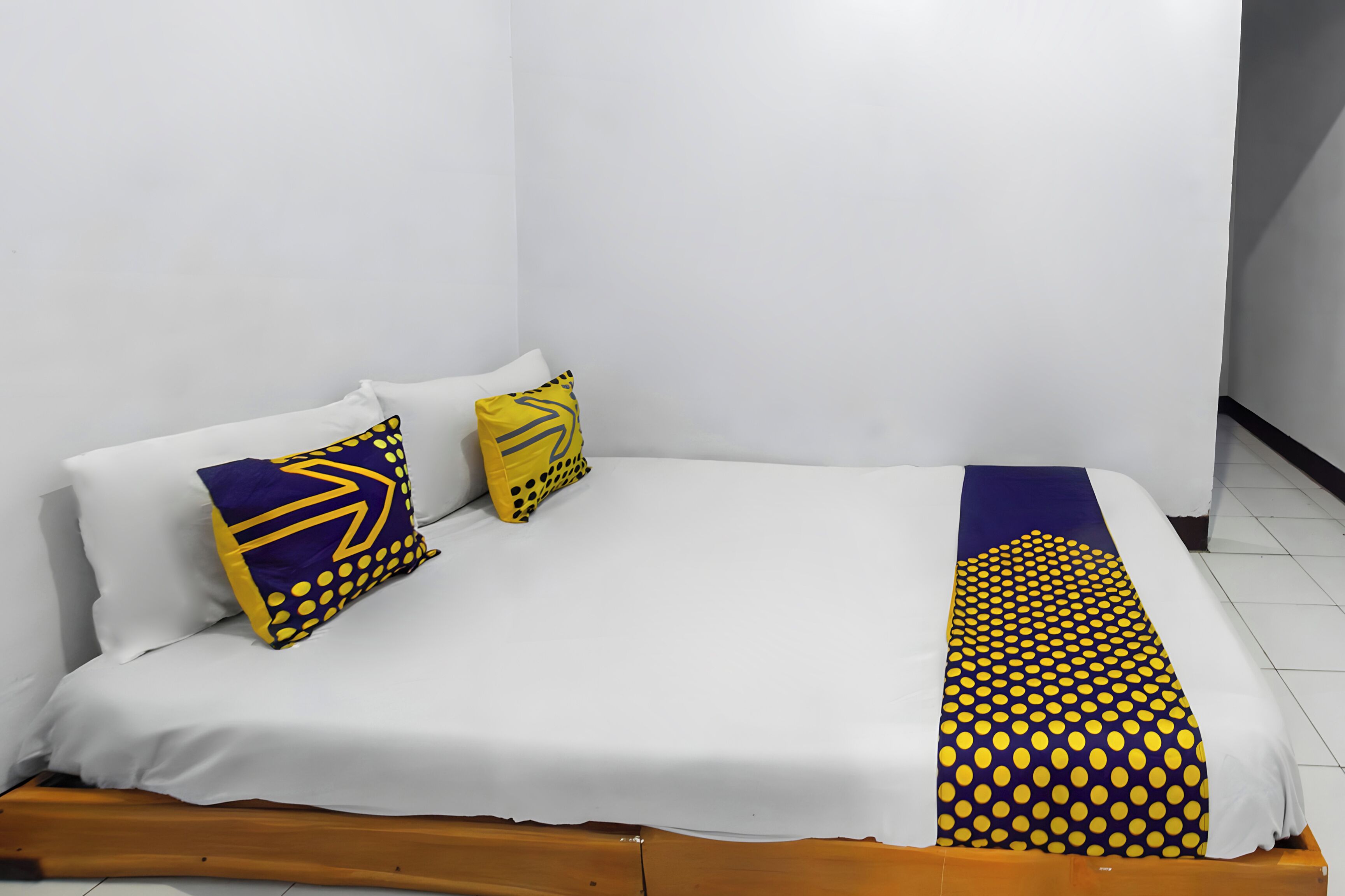 Una cama individual con ropa blanca, dos almohadas con diseños geométricos y un plaid amarillo y azul con lunares.