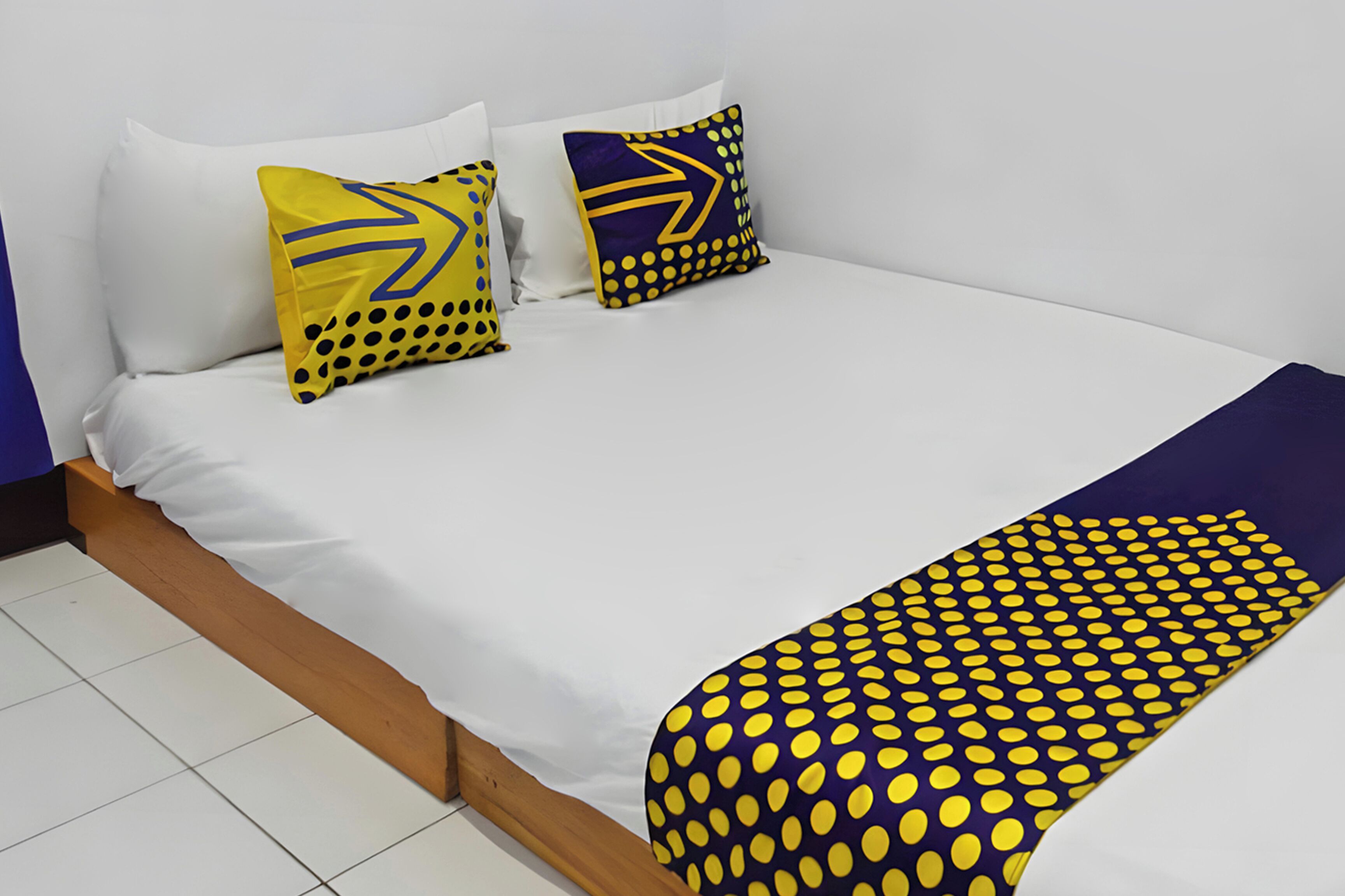 A bed with white bedding, two pillows with yellow and blue patterns, and a blue and yellow polka dot blanket.