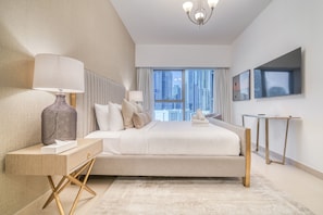 A modern hotel bedroom with a large bed, bedside tables, a desk, and a view of the cityscape through large windows.