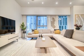 Un salon moderne doté d’un grand téléviseur à écran plat, d’un canapé beige, d’une table basse et offrant une vue sur la ville grâce à de grandes fenêtres.