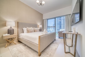 A modern bedroom with a large bed, bedside lamps, a nightstand, a desk, and a view of a cityscape through the window.