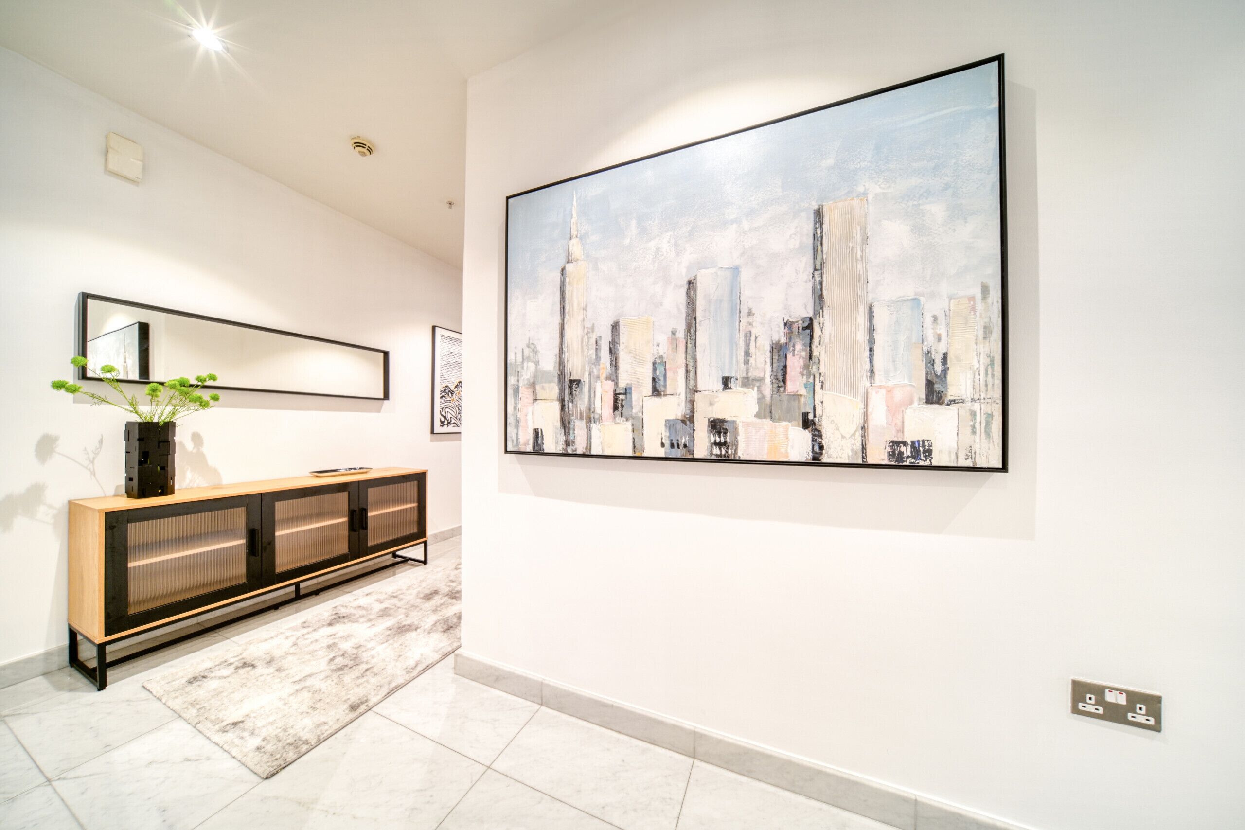 Un corridoio con un grande quadro astratto raffigurante un paesaggio urbano, una consolle in legno con ante in vetro, uno specchio e un vaso con piante verdi.