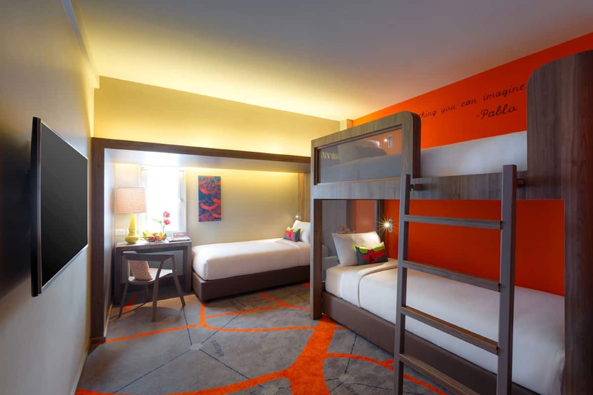 A hotel room with a bunk bed, a desk with a chair, a lamp, and a wall with a quote.