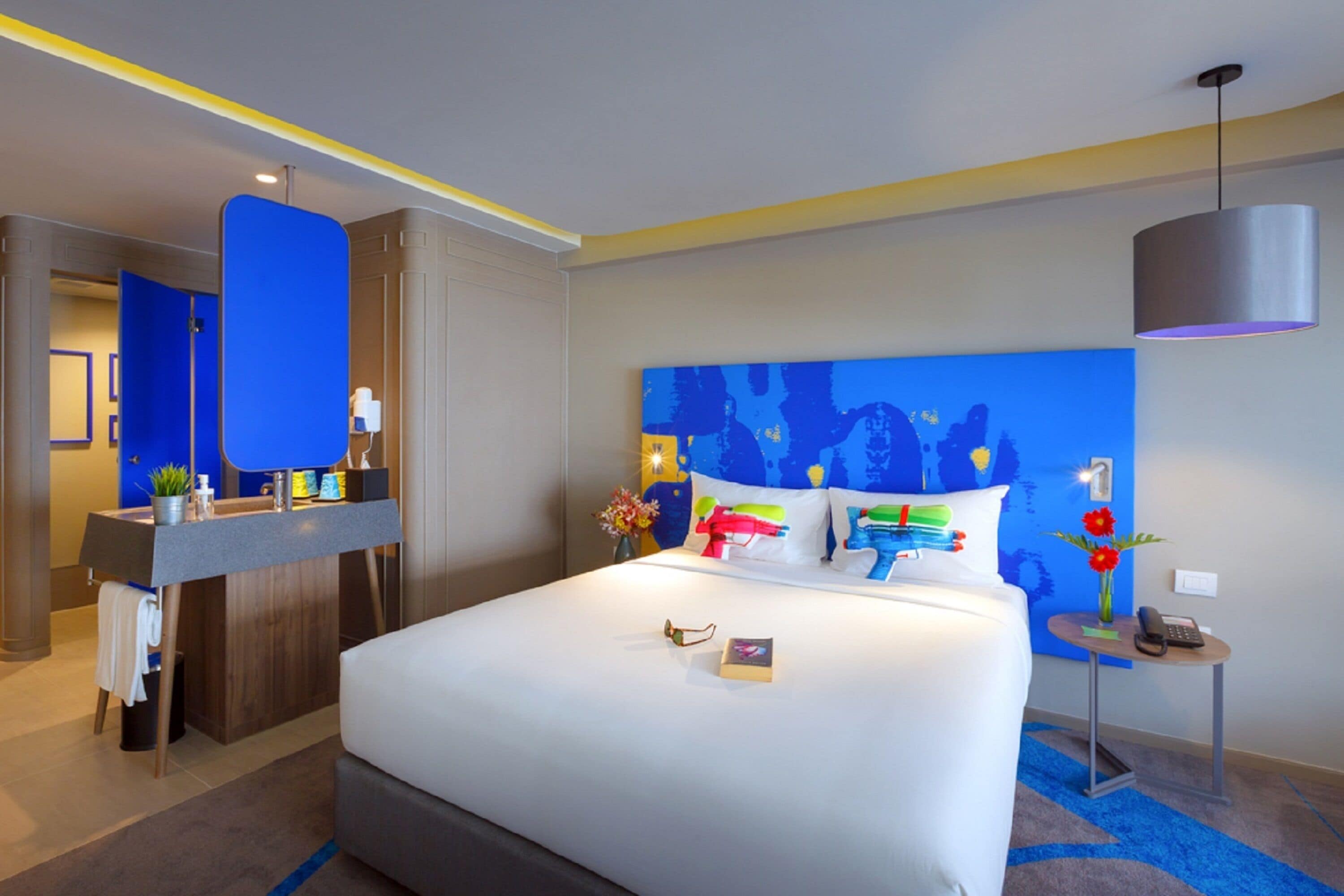 A modern hotel room with a large bed, a bedside table, a desk with a chair, and a bathroom visible through a door.