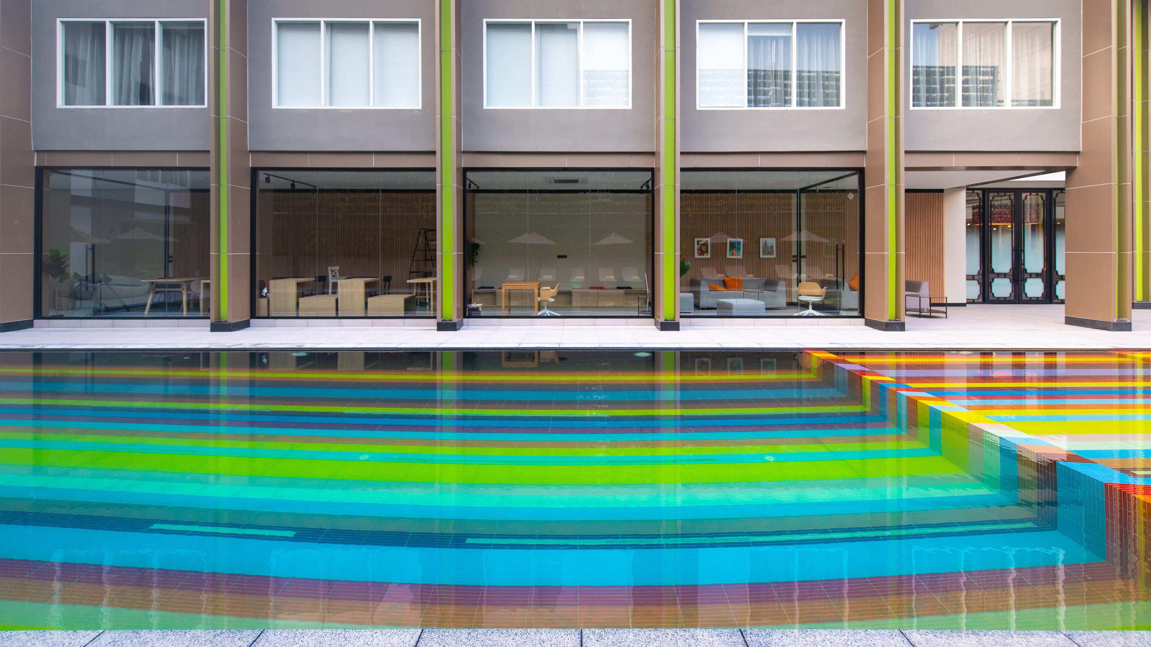 A modern building with a glass facade, a pool with colorful steps, and a seating area with tables and chairs.