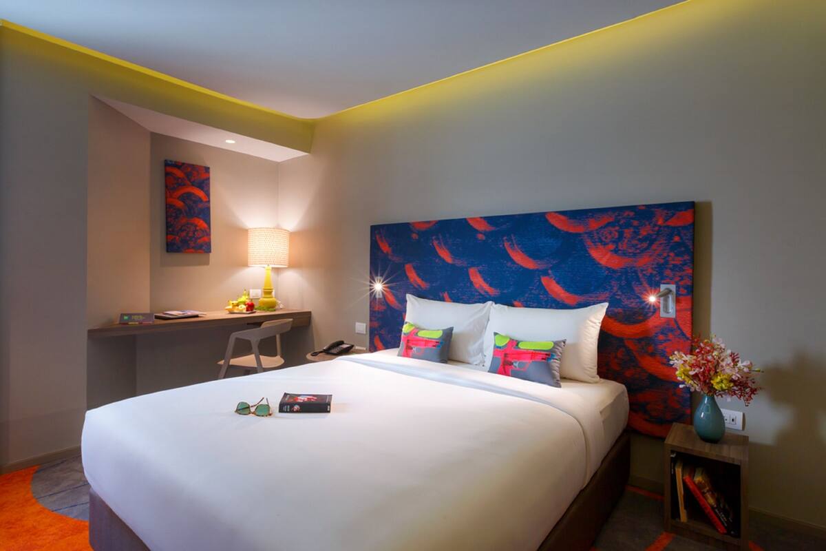 A hotel room with a bed, a desk, a chair, a lamp, and a nightstand with a vase.