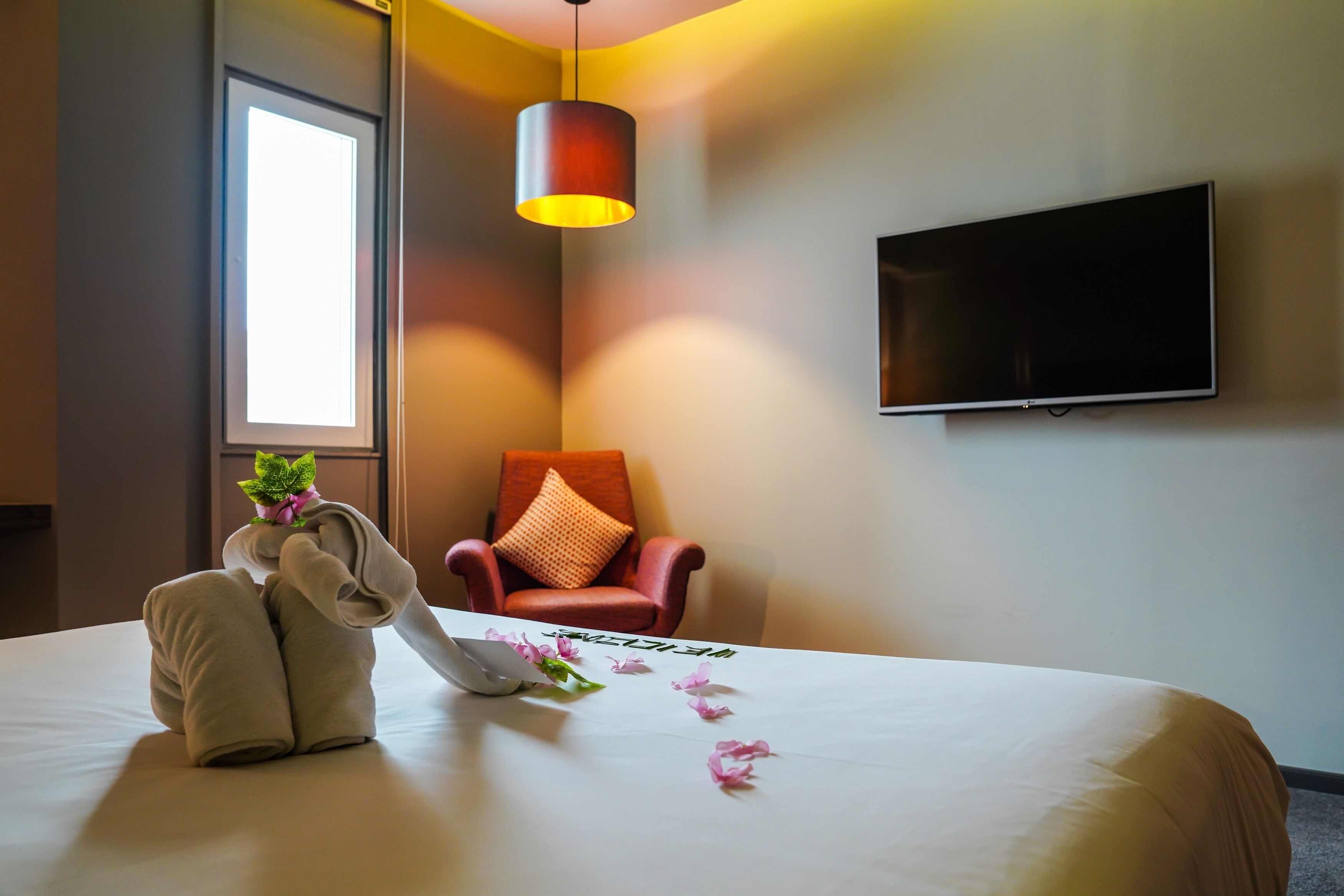A hotel room with a bed, a TV mounted on the wall, a chair with a cushion, and a window providing natural light.
