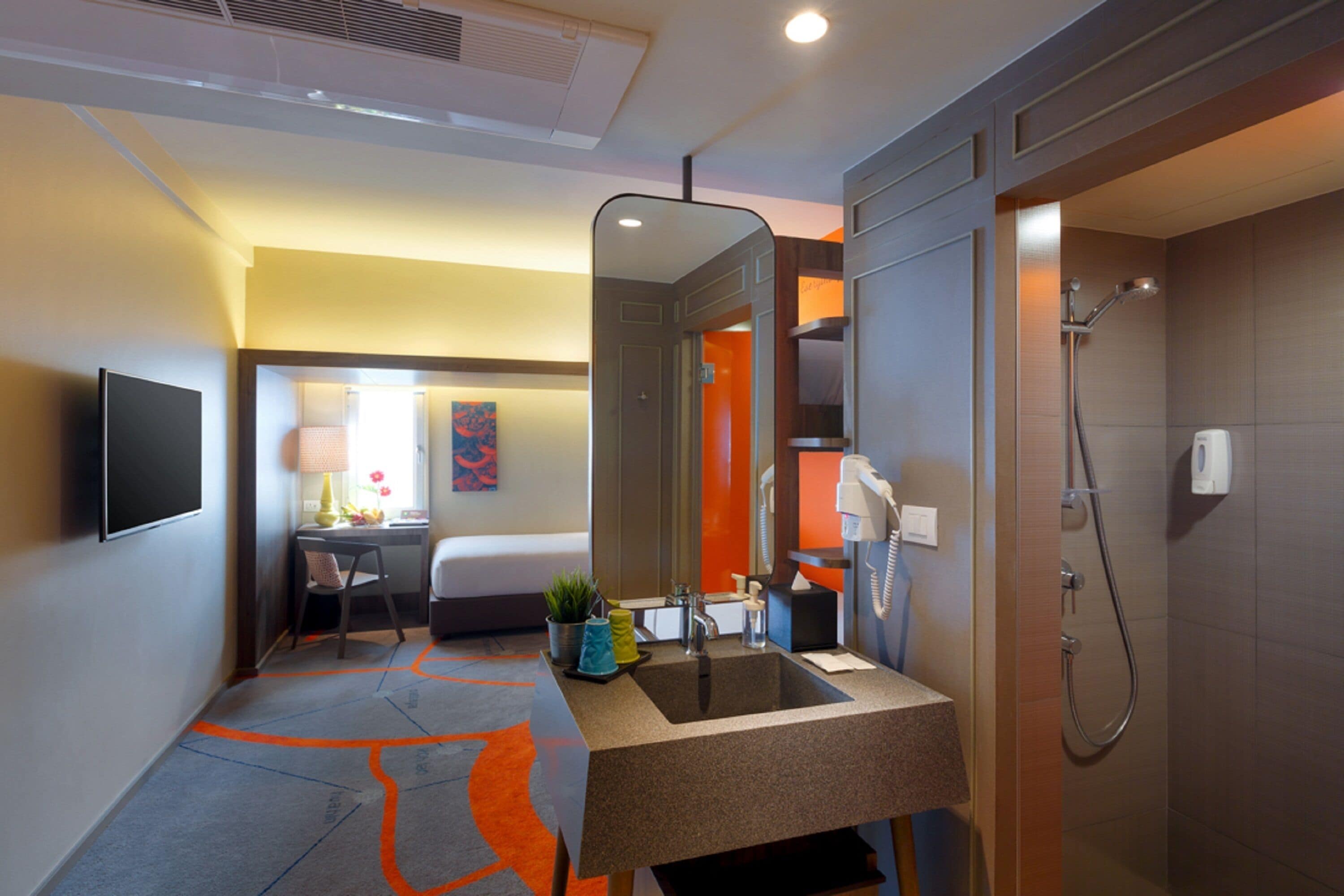 Bagno d'albergo con lavandino, specchio, doccia e un letto sullo sfondo.