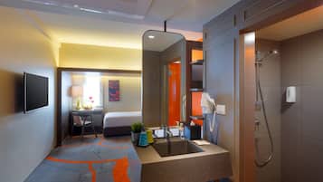 Baño de hotel con lavamanos, espejo, ducha y cama al fondo.