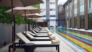 Un área junto a la piscina con sillones de descanso y sombrillas, un edificio moderno al fondo y un deck de piscina colorido.