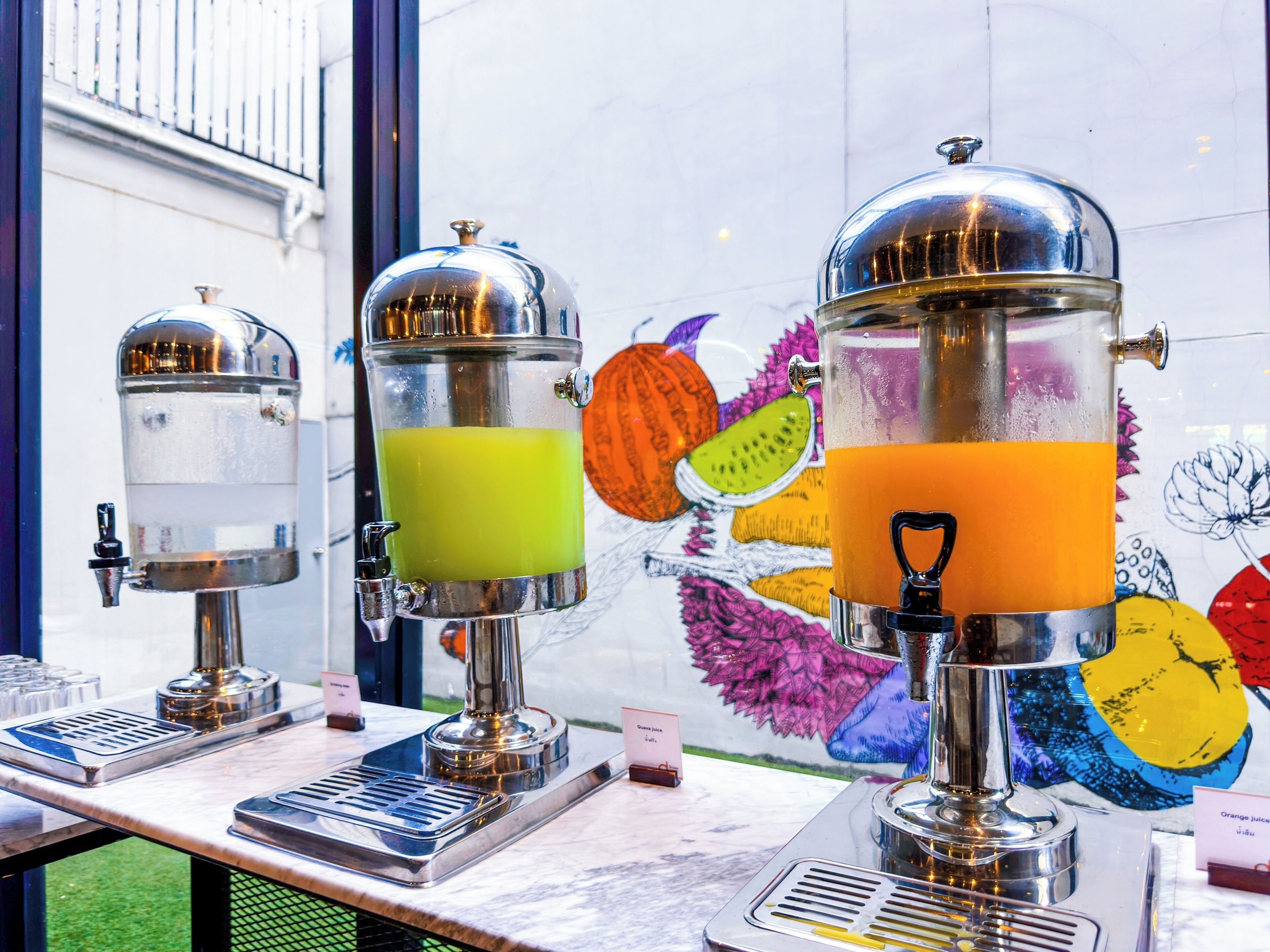 A juice bar with two stainless steel dispensers, one with green juice and the other with orange juice, against a wall with a colorful mural.