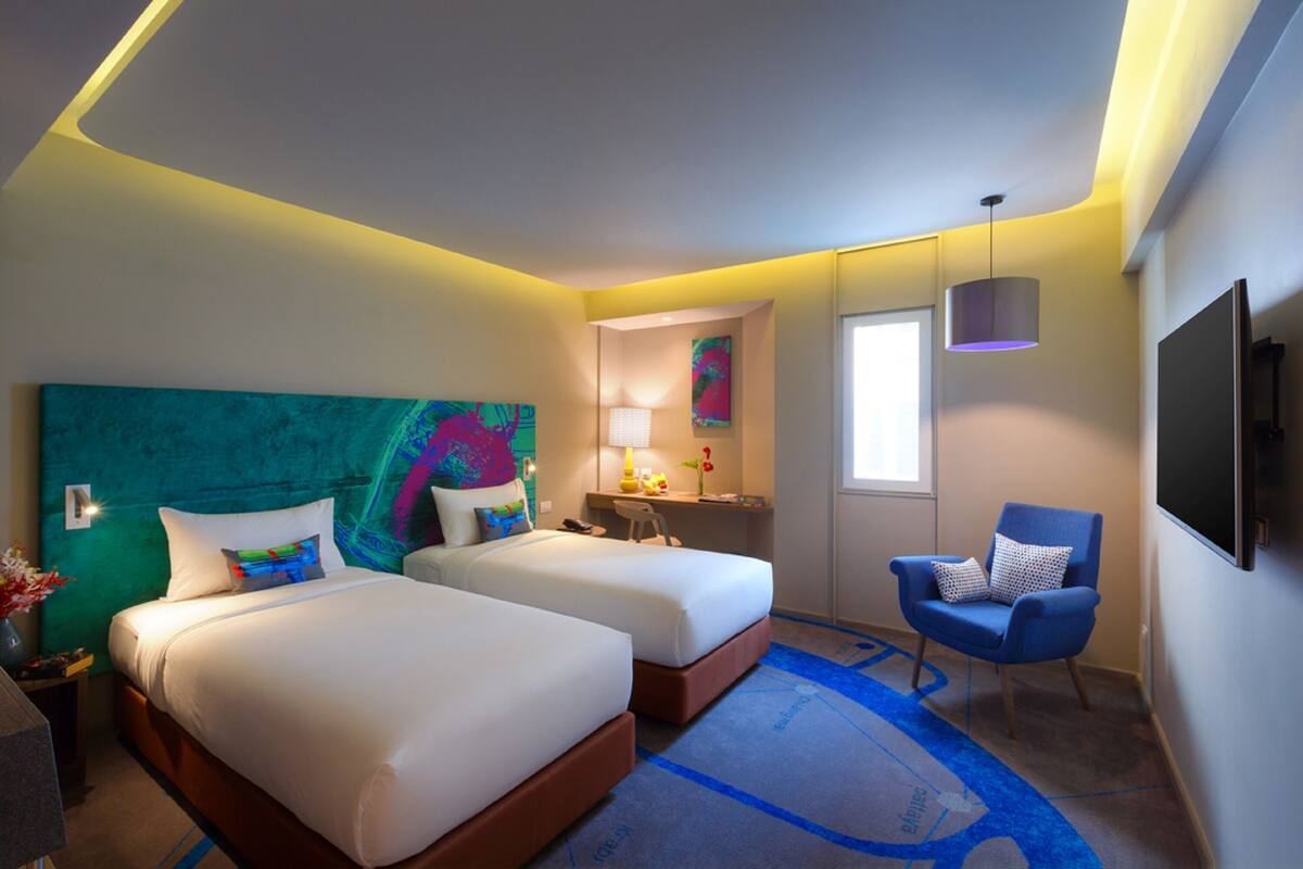A hotel room with two beds, a desk, a chair, a TV, and a large abstract painting on the wall.