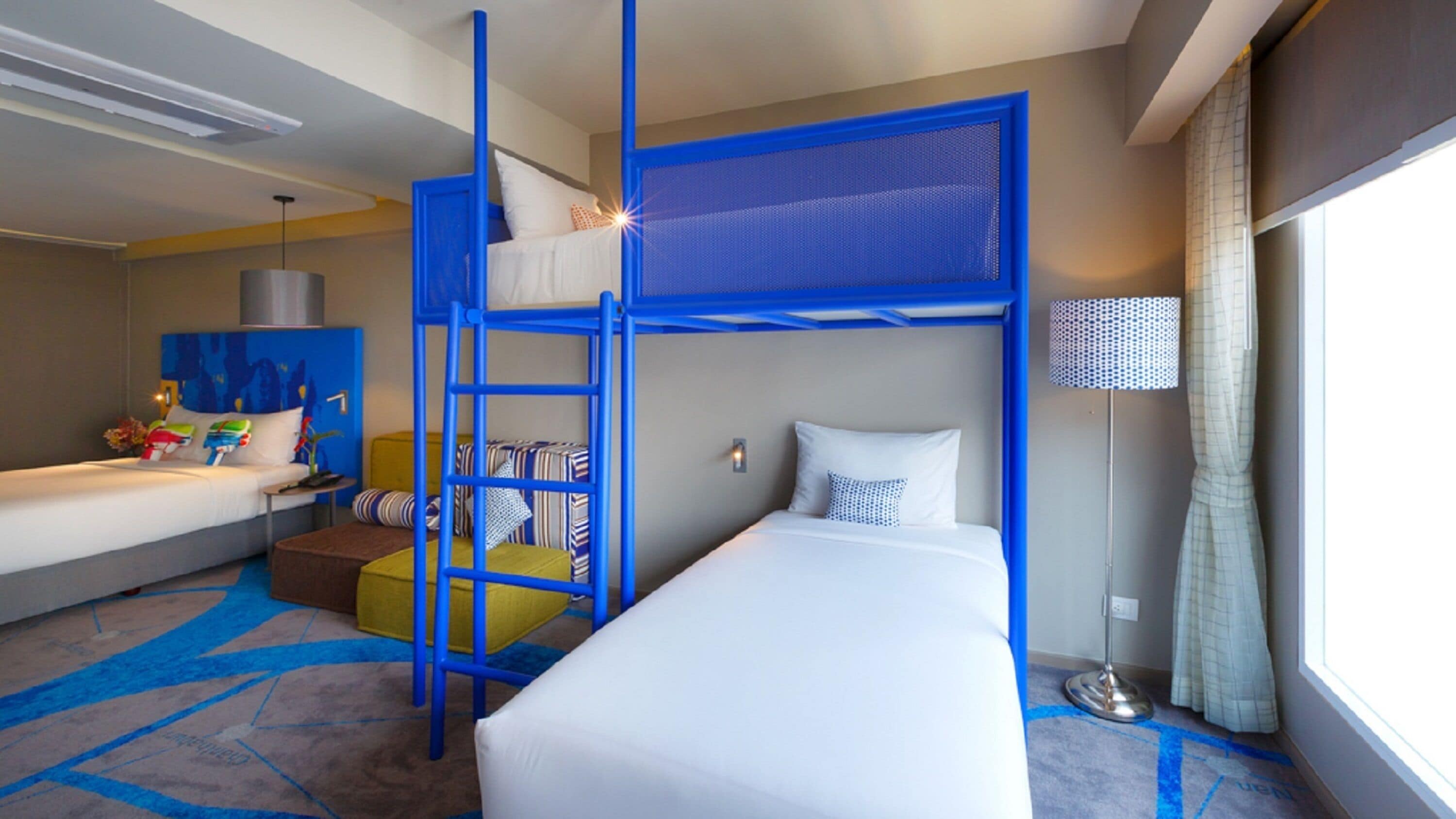 A hotel room with a bunk bed, a single bed, a small seating area, and a window with curtains.