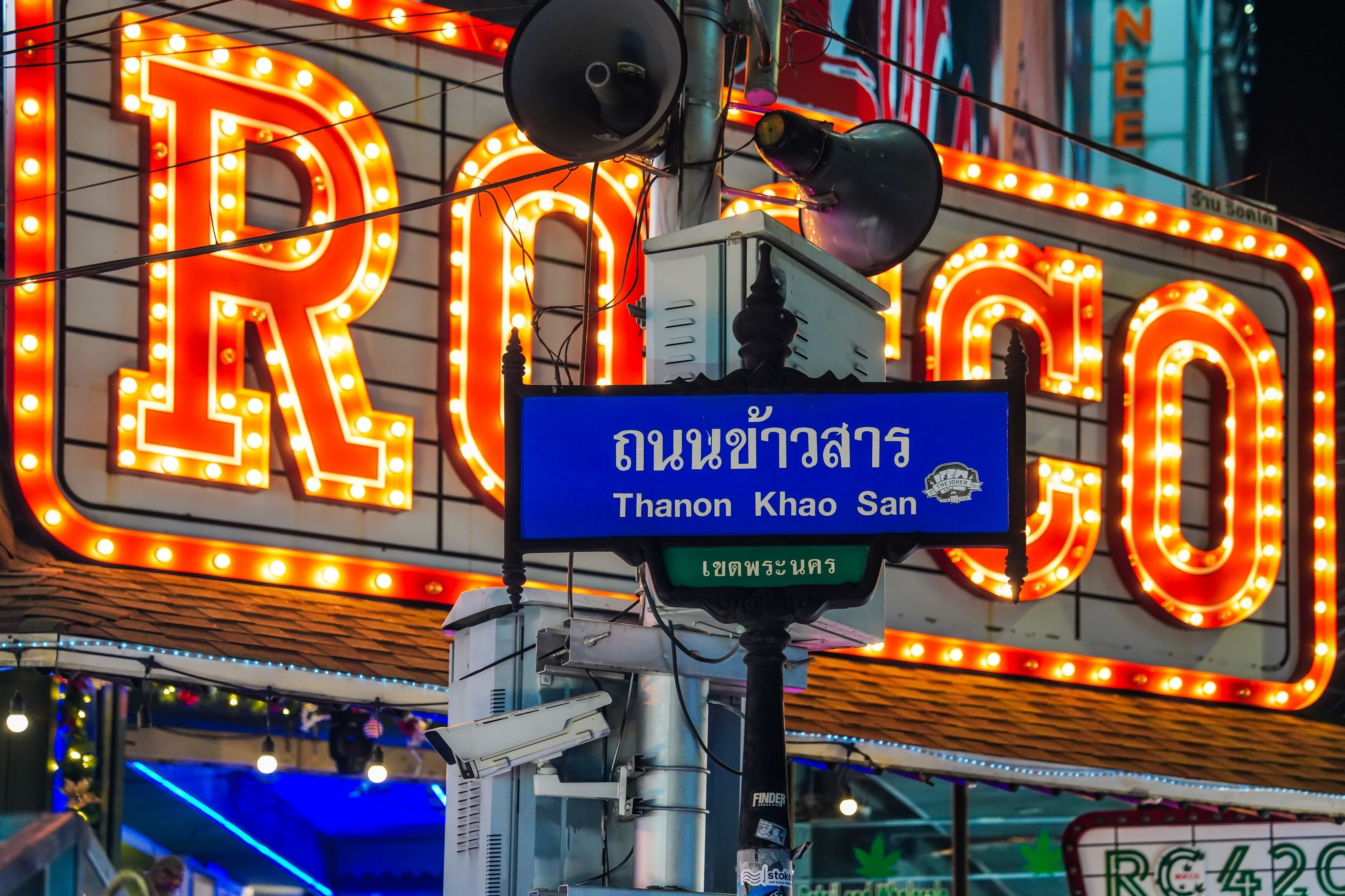 Une enseigne au néon avec les lettres « R » et « O » illuminées en rouge, et une enseigne bleue en dessous avec du texte en thaï.