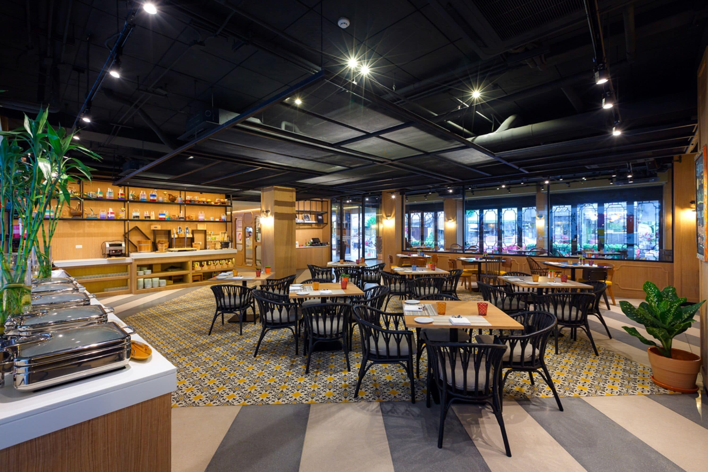 Un restaurant doté de nombreuses tables et chaises, d’un espace buffet et de grandes fenêtres offrant une vue sur la ville.