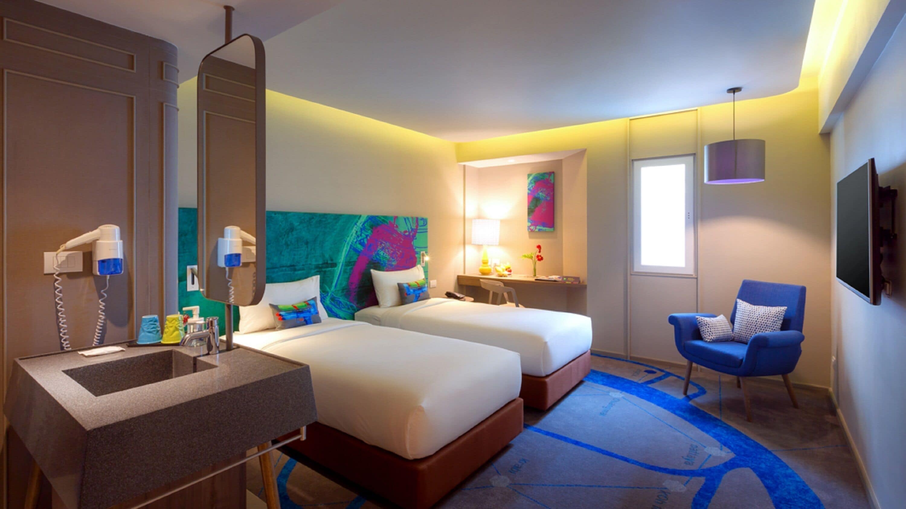 Habitación de hotel con dos camas, baño con lavamanos y espejo, una silla y un cuadro en la pared.
